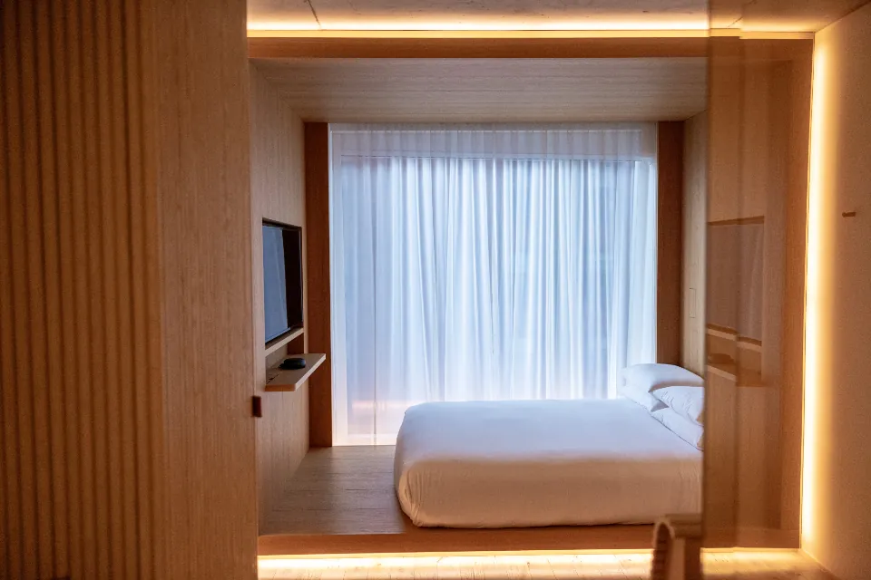 Bedroom lined in pale timber joinery, with a lit alcove framing the bed and a sheer curtain beyond