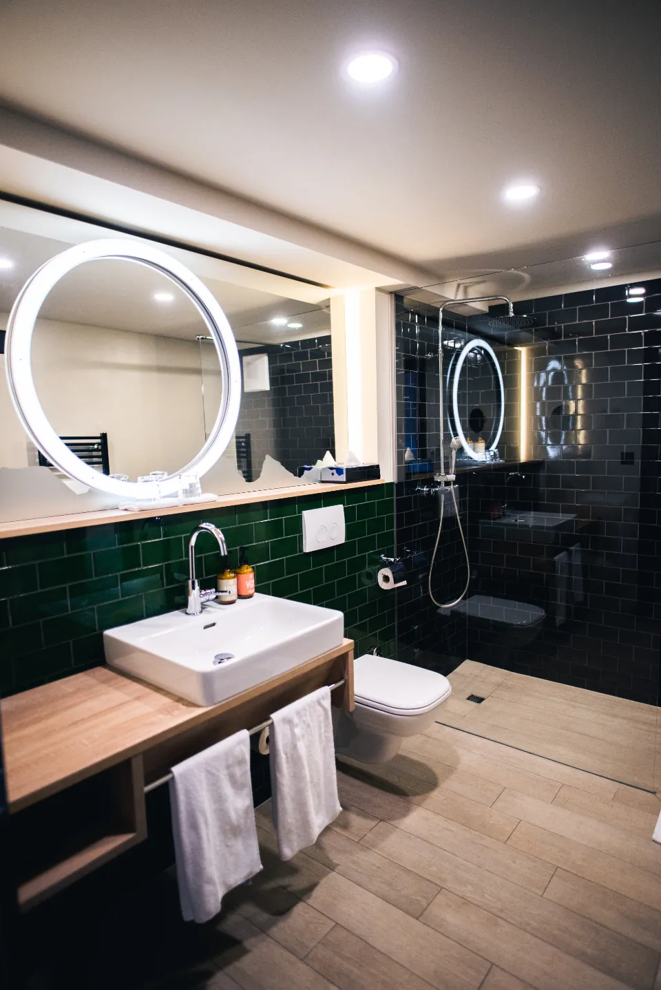 Bathroom with green wall tiles, a timber vanity and a round lit mirror